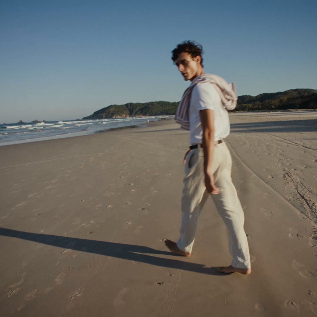 A young man walks along a sandy beach with the ocean on his left and rolling green hills in the distance. He is wearing a white t-shirt and light-colored linen pants, with a striped towel draped over his shoulders. The sun is low in the sky, casting a long shadow behind him. Text overlays the image, reading "Part memories, part moments. Familiar, effortless and distinctly Australian."