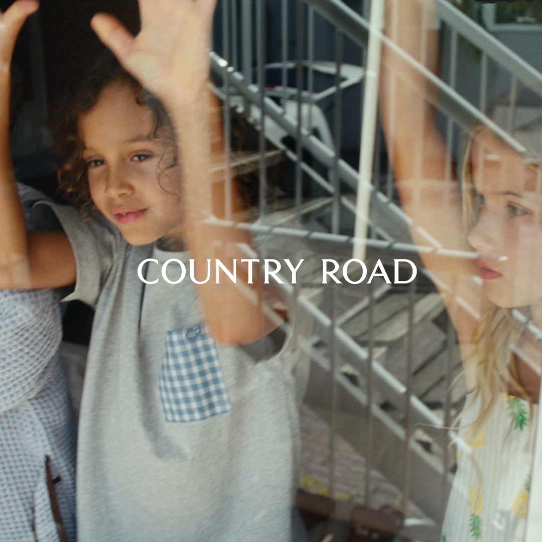 Two young children, a boy and a girl, are looking out of a window, their hands pressed against the glass. The boy in the foreground has curly brown hair and is wearing a grey t-shirt with a blue checkered pocket. The girl on the right has blonde hair and is wearing a white top with a pineapple print. White text overlaid on the image reads "COUNTRY ROAD". The overall scene evokes a sense of childhood innocence and perhaps longing, with the children appearing to be looking out at something or someone.