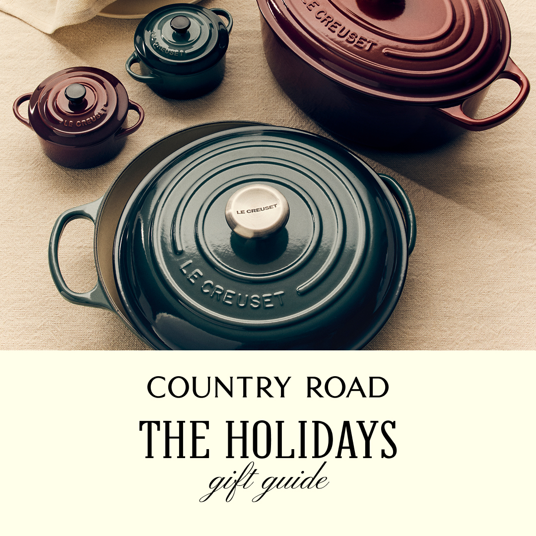 A top-down view of several Le Creuset cast iron pots and pans in deep green and burgundy colors, arranged on a textured beige tablecloth. The pots are in various sizes, with lids on most of them. The bottom portion of the image features a cream-colored background with text overlay. The text reads "COUNTRY ROAD" in a simple serif font, followed by "THE HOLIDAYS" in a larger, bold serif font, and "gift guide" in an elegant script font below it. The overall aesthetic is warm and inviting, suggesting a holiday gift guide.