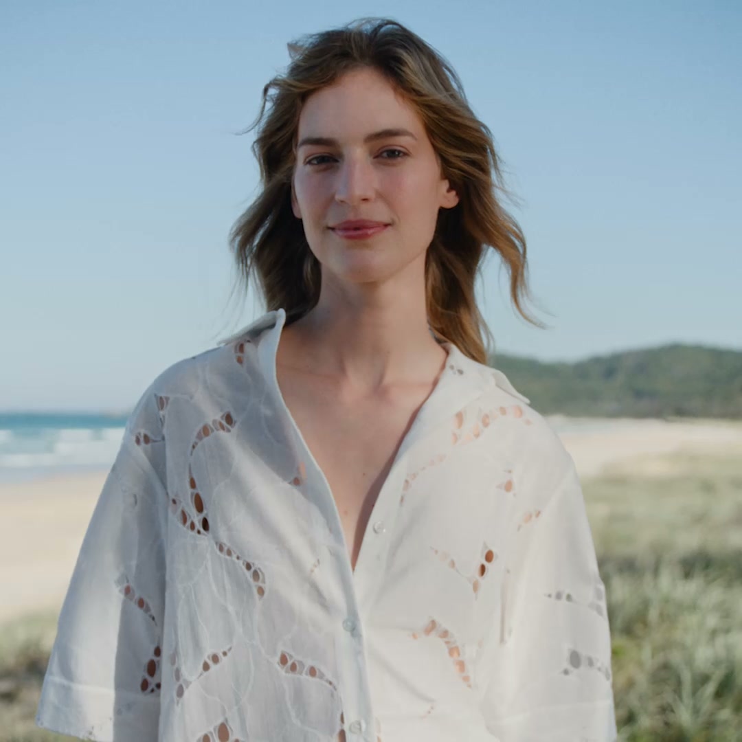 A woman stands on a beach with the ocean and dunes in the background. She is wearing a white, open-collared shirt with intricate cut-out details. The text "Still on your mind?" is overlaid on the image. The ad copy suggests shopping for occasionwear, summer accessories, and everyday essentials.