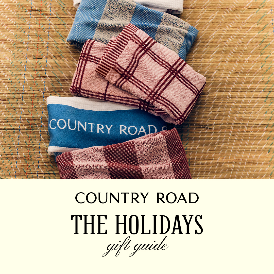 A stack of folded towels in various plaid patterns and colors are laid out on a woven mat. One towel is blue with white text that reads "COUNTRY ROAD". Below the towels, overlaid on a cream background, is text that reads "COUNTRY ROAD" in a simple font, followed by "THE HOLIDAYS" in a larger, bold serif font, and then "gift guide" in a script font. The overall impression is a curated collection of items suitable for holiday gifting.
