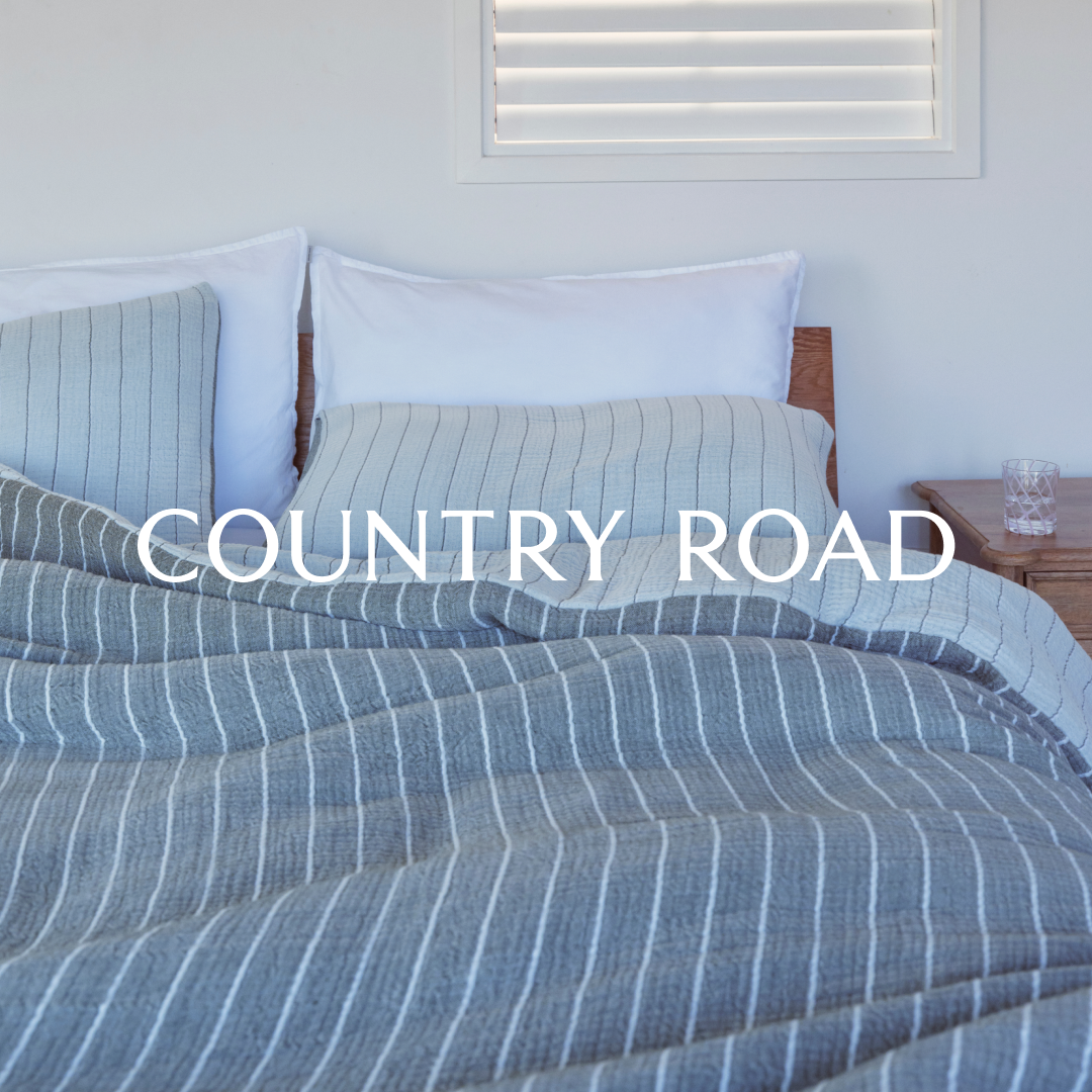 A neatly made bed with white sheets and pillows is shown, covered by a grey and white striped duvet. The brand name "COUNTRY ROAD" is overlaid in large white sans-serif font across the center of the image. The scene is bright and airy, suggesting a bedroom setting with a bedside table and a glass of water visible on the right. The overall aesthetic is clean and minimalist, focusing on the bedding and the brand's identity.