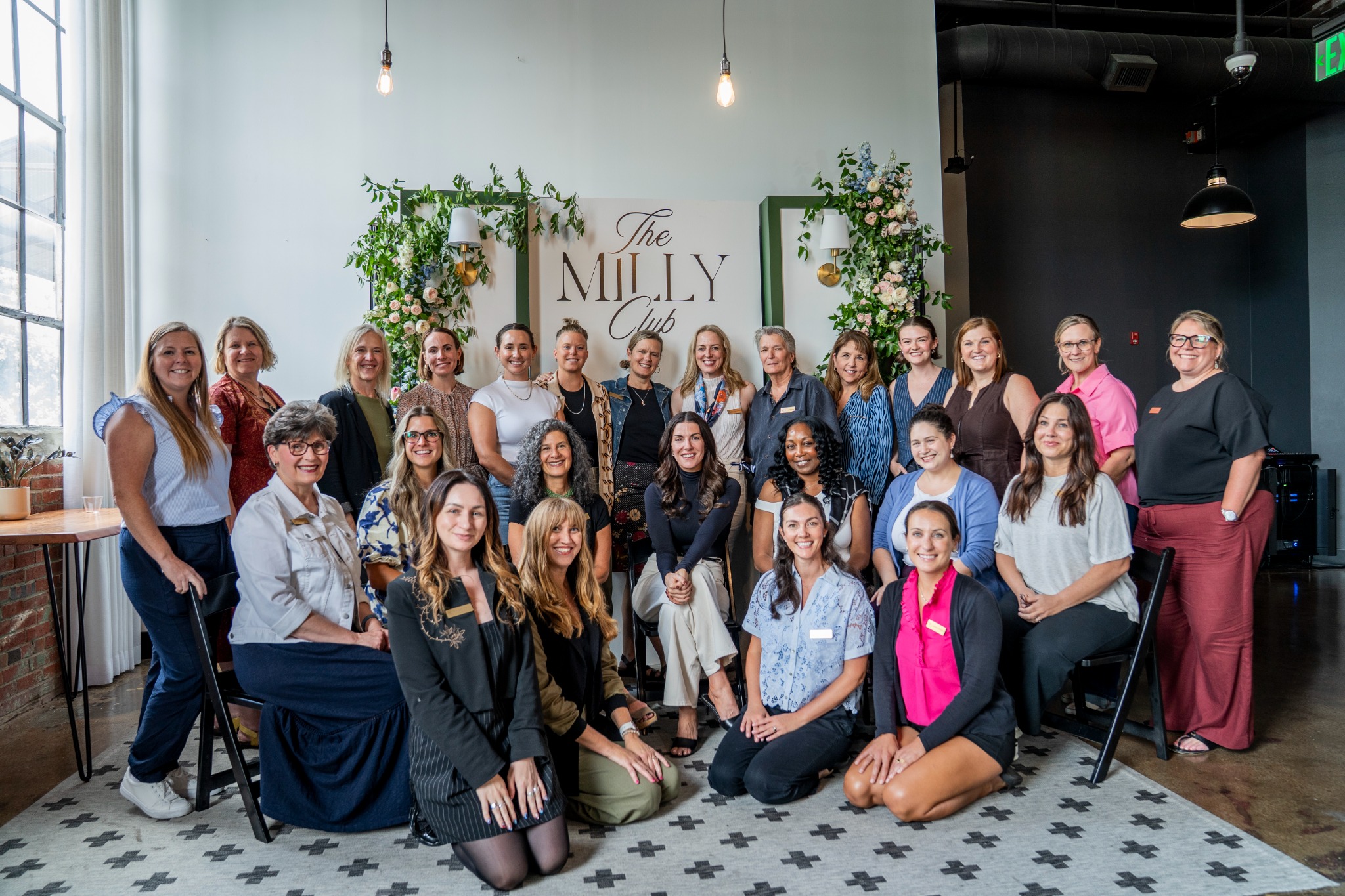 A diverse group of approximately 25 women are posing for a group photo in an indoor setting. The women are of various ages and ethnicities, and they are dressed in casual to business-casual attire. Some are standing, while others are seated or kneeling in the front row. Behind them, a large sign reads "The Milly Club" in elegant script. The overall atmosphere appears to be one of community and connection, with many of the women smiling warmly at the camera. The text "We're all looking for our people." is overlaid on the image.