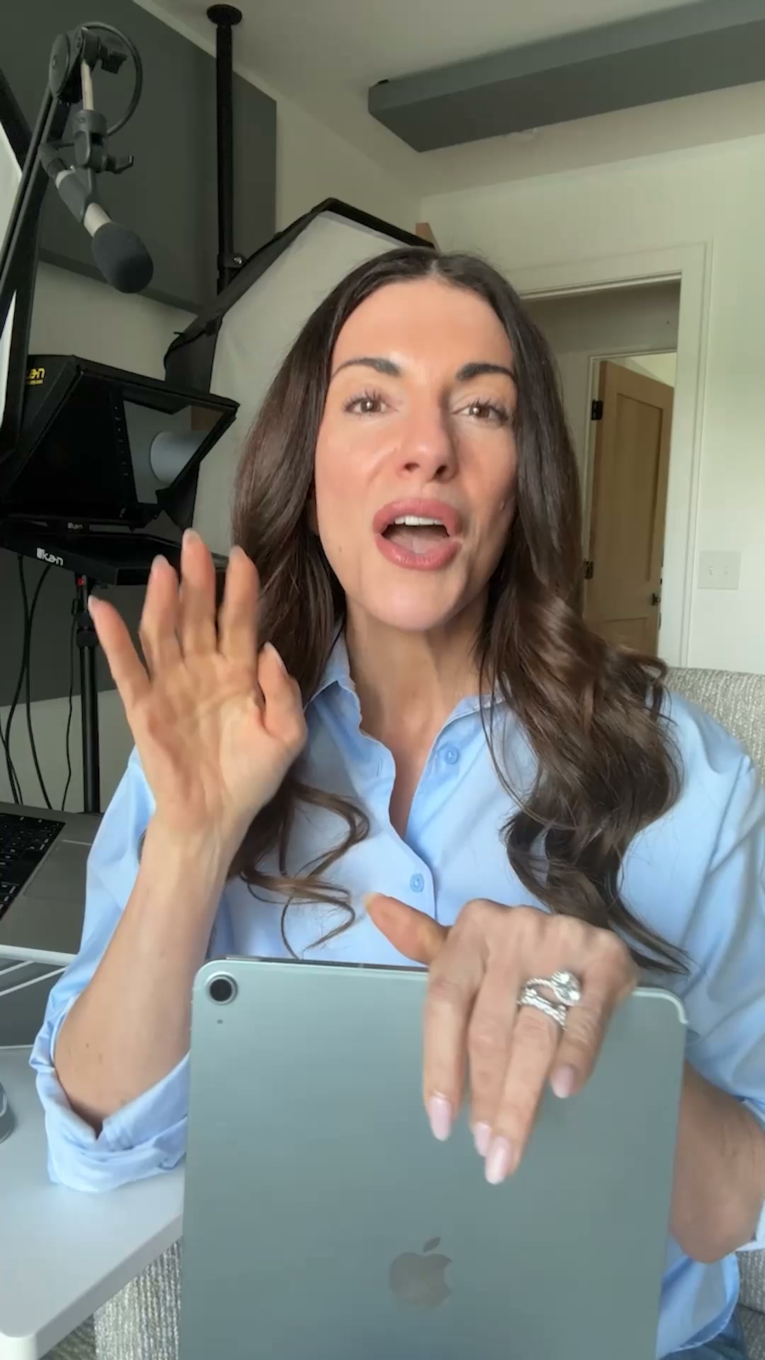 A woman with long, dark, wavy hair, wearing a light blue button-down shirt, is speaking directly to the camera. She is holding a light blue tablet in her left hand and gesturing with her right hand. The background shows a home office setup with a ring light and a teleprompter. Text overlays appear on the screen, detailing the contents of 'The Milly Club' mastermind program, including a business scaling framework, accountability, direct communication, expert guidance from a $130M founder, and in-person events. The hook is a question posed directly to the viewer about their decision regarding the club.