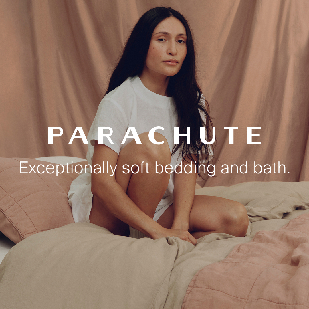 A woman with long dark hair is sitting on a bed, wearing a white t-shirt and shorts. She is looking directly at the camera with a neutral expression. The background is a soft, draped fabric in a muted rose color. Large white text on the image reads "PARACHUTE" and below it, "Exceptionally soft bedding and bath." The bedding is in muted tones of pink and beige.