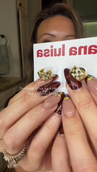 A woman's hands are shown holding a white card with red text that reads "siul 6" backwards. She is also holding two gold hoop earrings, one encrusted with diamonds and the other with a checkered pattern of diamonds. The text "Nothing cures looking frumpy like a classic hoop" is overlaid on the image. The woman has long, almond-shaped nails with a dark brown and nude tortoiseshell design, and small gold star embellishments on some nails. A large diamond ring is visible on her left hand.