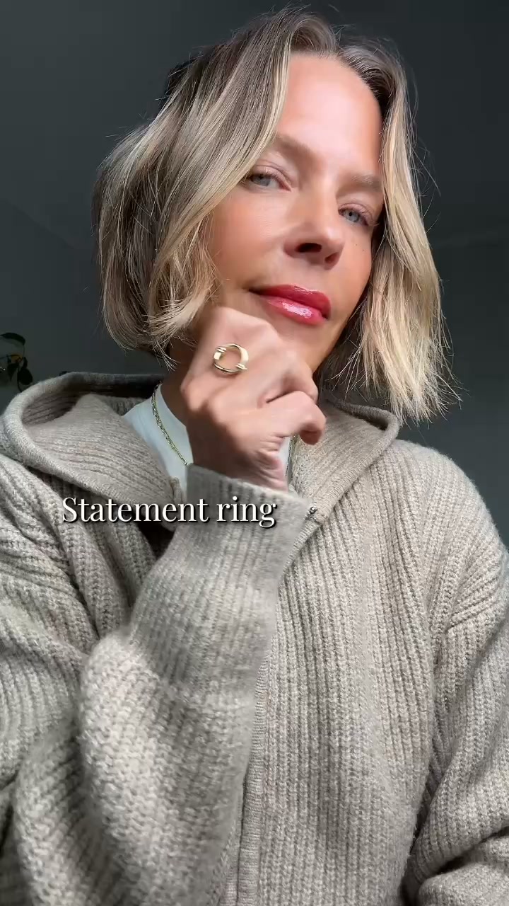 A woman with blonde, shoulder-length hair and red lipstick is shown from the chest up, wearing a beige knit hooded sweater. She is looking towards the camera with her hand resting on her chin, showcasing a gold statement ring on her index finger. The text "Statement ring" is overlaid on the image in a white sans-serif font with a black outline.