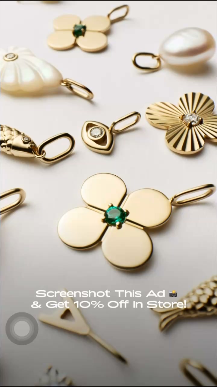 A close-up, overhead shot displays an assortment of gold pendants with various designs, including floral shapes, a pearl, and a shell. One prominent pendant in the foreground features a four-petal flower design with a green emerald at its center. White text overlaid on the bottom of the image reads "Screenshot This Ad & Get 10% Off In Store!" with a camera icon next to the word "Ad".