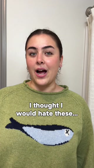 A young woman with dark hair and bold eyebrows, wearing a green sweater with a navy blue fish design, looks directly at the camera with an open mouth. White text overlaid on the sweater reads, "I thought I would hate these...". The background is a plain wall with a curtain on the right side. The creative appears to be a personal video from a creator sharing an initial negative opinion about a product.