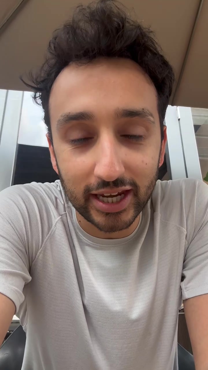 A man with dark curly hair and stubble is looking down and speaking. He is wearing a light grey t-shirt. The text "Introducing The Lifestyle Business Academy" is displayed at the top of the screen, and below it, "👉 A Roadmap to $100k/year" is shown. The creative is a close-up selfie-style video of the creator.