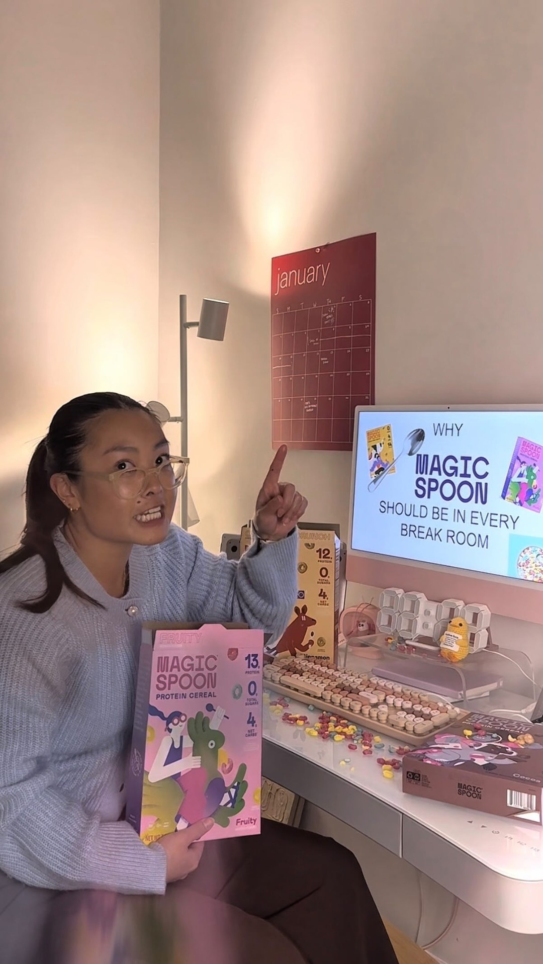 A woman with her hair in a ponytail, wearing glasses and a light blue sweater, holds a box of Magic Spoon cereal. She is sitting at a desk with a computer displaying text that reads "WHY MAGIC SPOON SHOULD BE IN EVERY BREAK ROOM". A calendar showing "January" is visible on the wall behind her. She is pointing her finger upwards, looking towards the camera with an engaging expression.