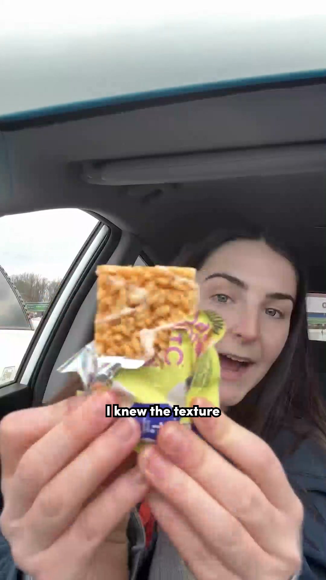 A young woman is holding up a Magic Spoon cereal bar to the camera, which she is about to eat. The bar is square and appears to be made of puffed rice and nuts. The woman is in a car, and the background shows trees and a cloudy sky. Text on the screen reads "I knew the texture." The woman is smiling and looking at the camera.