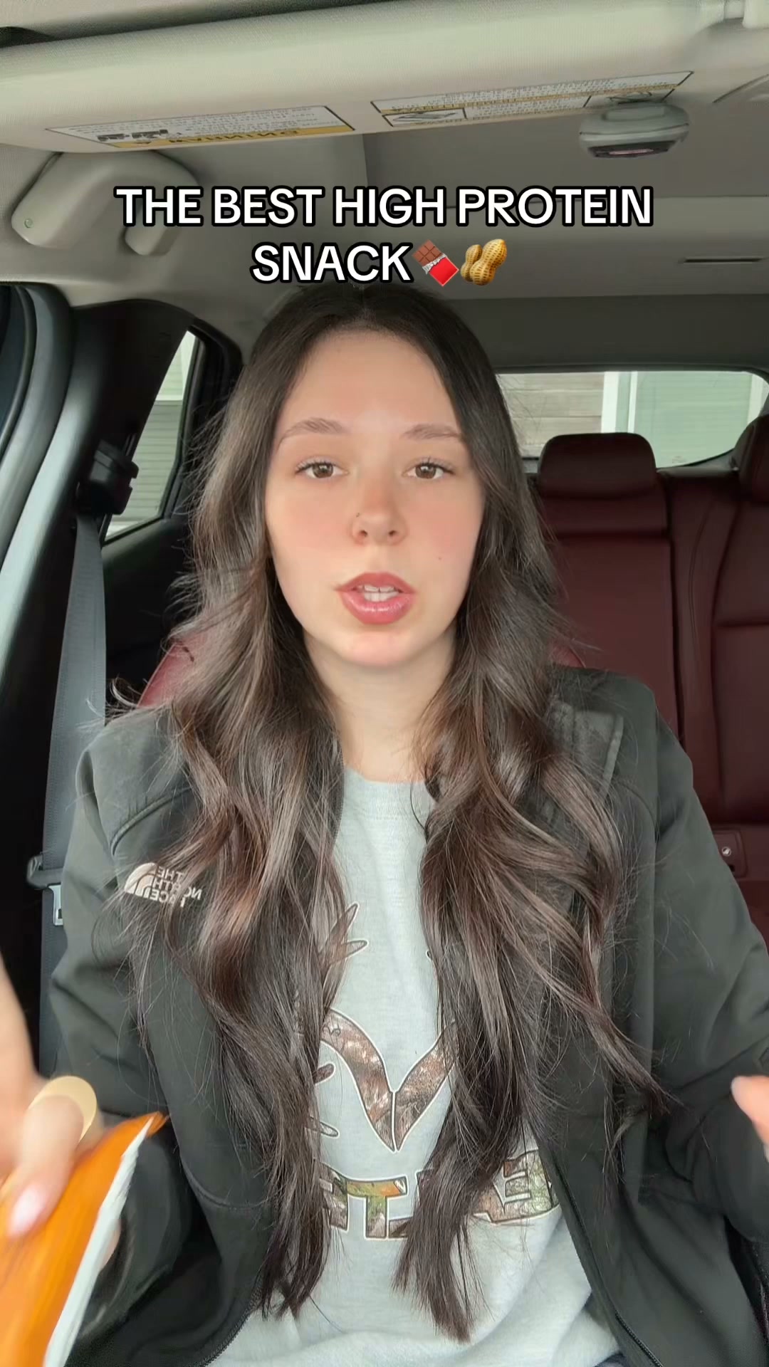 A young woman with long, dark, wavy hair is sitting in the driver's seat of a car. She is wearing a black jacket over a grey t-shirt with a camouflage deer logo. White text overlaying the top of the image reads "THE BEST HIGH PROTEIN SNACK" with a chocolate bar and peanut emoji. She is looking directly at the camera with her mouth slightly open, as if speaking. In the lower left corner, a hand is holding an orange and white snack bar. The background shows the car's interior, including the headrest of the seat behind her.