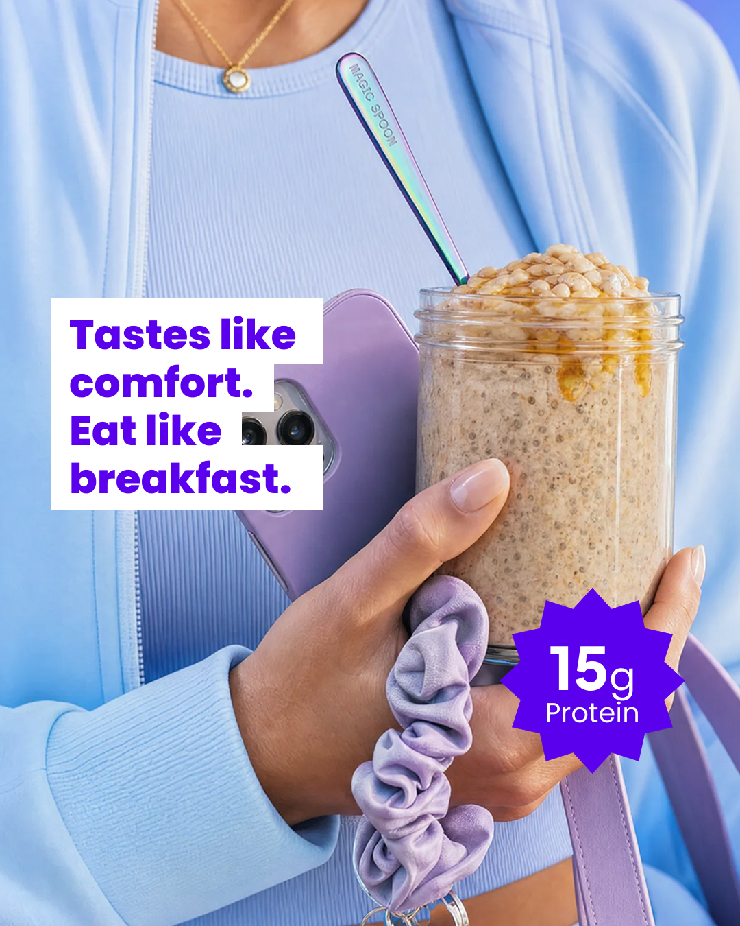 A close-up shot shows a person holding a jar of overnight oats and a purple smartphone. The jar is topped with honey and what appears to be granola or nuts. A spoon with "MAGIC SPOON" printed on it is in the jar. Text overlays the image in bold purple and white font, reading "Tastes like comfort. Eat like breakfast." A purple starburst graphic in the lower right corner highlights "15g Protein."