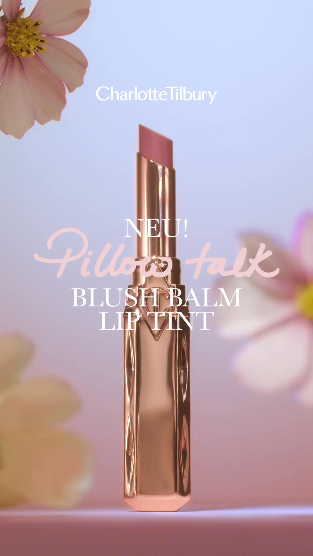 A close-up shot of a rose-gold lipstick tube with the cap off, revealing a pink lipstick bullet. The Charlotte Tilbury logo is visible at the top. Text overlays read "NEU!", "Pillow talk", and "BLUSH BALM LIP TINT". Soft pink and yellow flowers are blurred in the foreground and background, creating a dreamy aesthetic. The overall impression is of a luxurious beauty product.