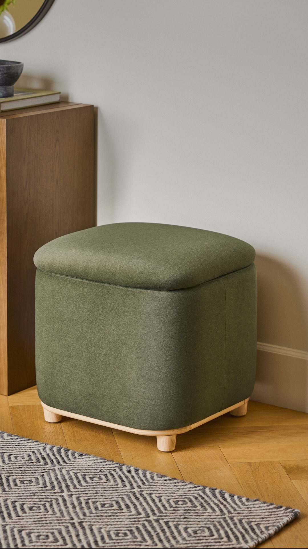 A square, olive-green storage ottoman with a rounded top sits on a wooden floor next to a dark wood cabinet. A patterned rug is visible in the foreground. The text "Less mess, more personality." is overlaid on the image, followed by "Our storage styles make it easy."