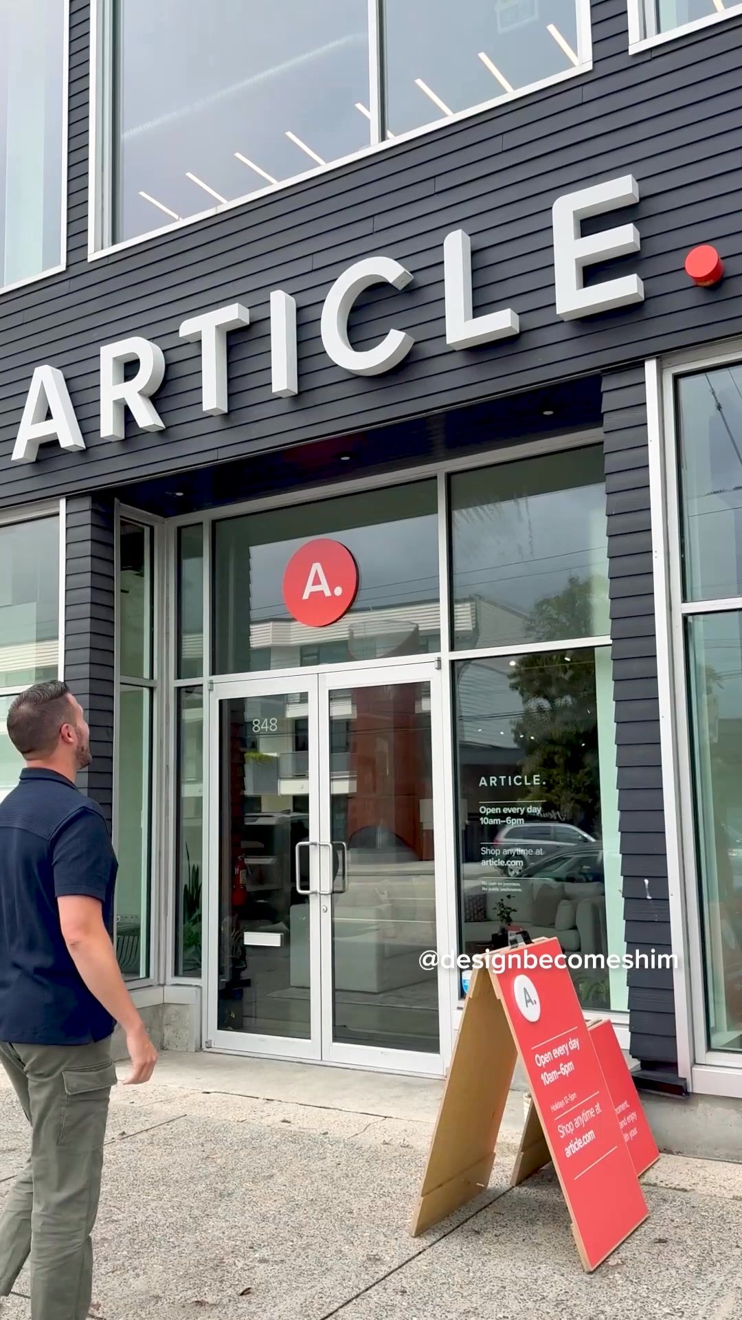 A man wearing a dark blue polo shirt and olive green cargo pants looks up at the exterior of the Article store. The store has large windows and prominent white "ARTICLE" lettering on a dark grey facade. A red circular logo with a white "A." is visible on the glass door. A red A-frame sign on the sidewalk displays store hours and website information. Text overlays on the image include the store's address and the creator's handle, "@designbecomeshim." The copy describes the creator's positive experience exploring the store and interacting with the staff.