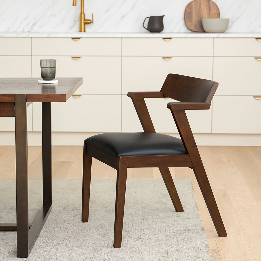 A modern dining chair with a dark wood frame and a black leather seat is positioned next to a dark wood dining table. A glass of water sits on the table. The background shows a clean, modern kitchen with cream-colored cabinets and a marble backsplash. White text overlaid on the image reads: "Great style is easy. From sofas to rugs, get your entire cart delivered with Article's $69 flat-rate shipping."