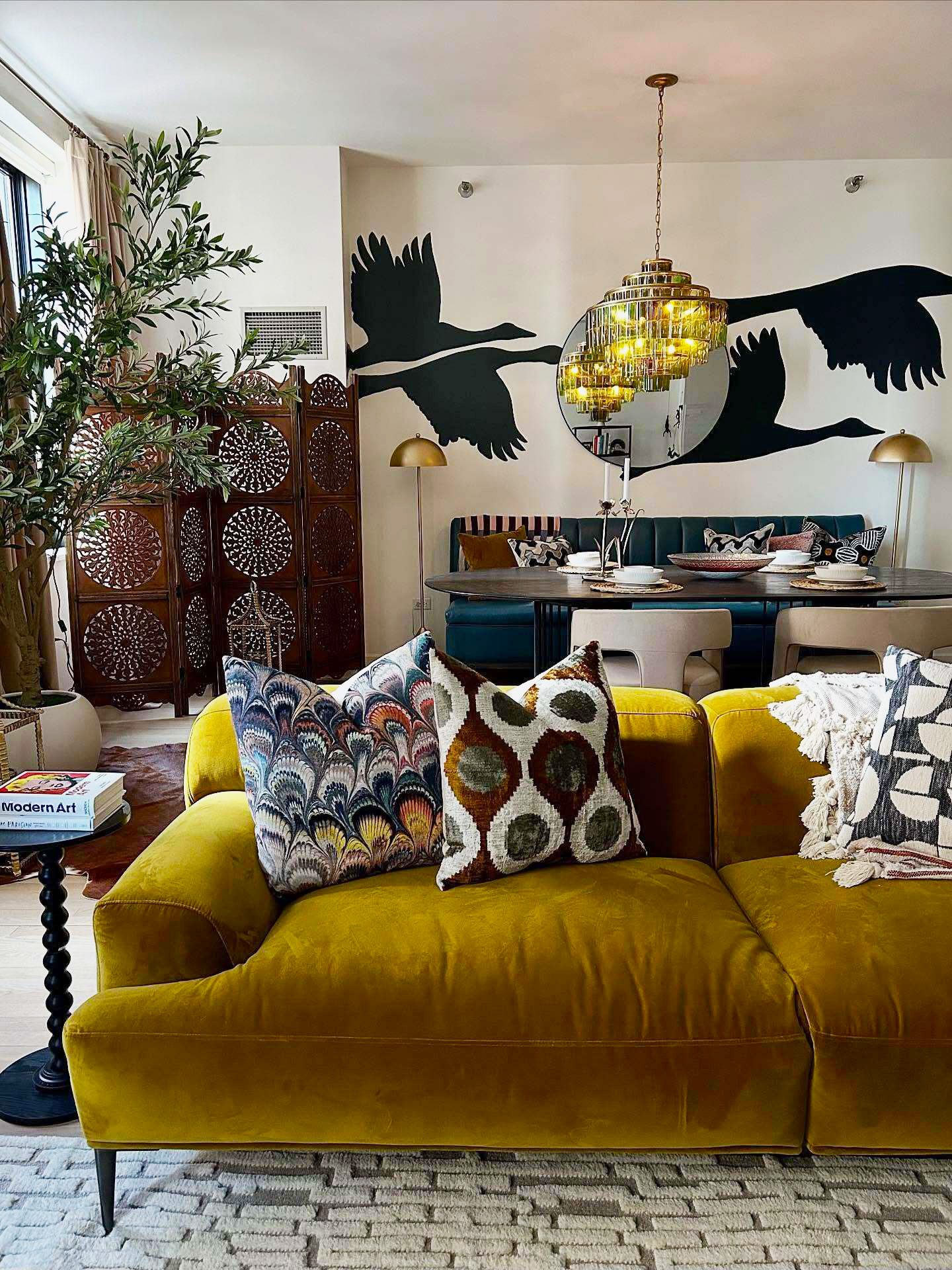 A richly decorated living room features a plush, mustard-yellow velvet sofa in the foreground. Behind the sofa, a dining area is visible with a dark, oval table set for a meal, a banquette, and chairs. The wall behind the dining area is adorned with a large, black silhouette mural of flying geese and a circular mirror reflecting the room. On-screen text overlays the image, stating 'Bold, golden, and made to glow ✨' and mentioning 'The Abisko Velvet Sofa in Plush Yarrow Gold' and crediting the photographer @mikeharrisonart.