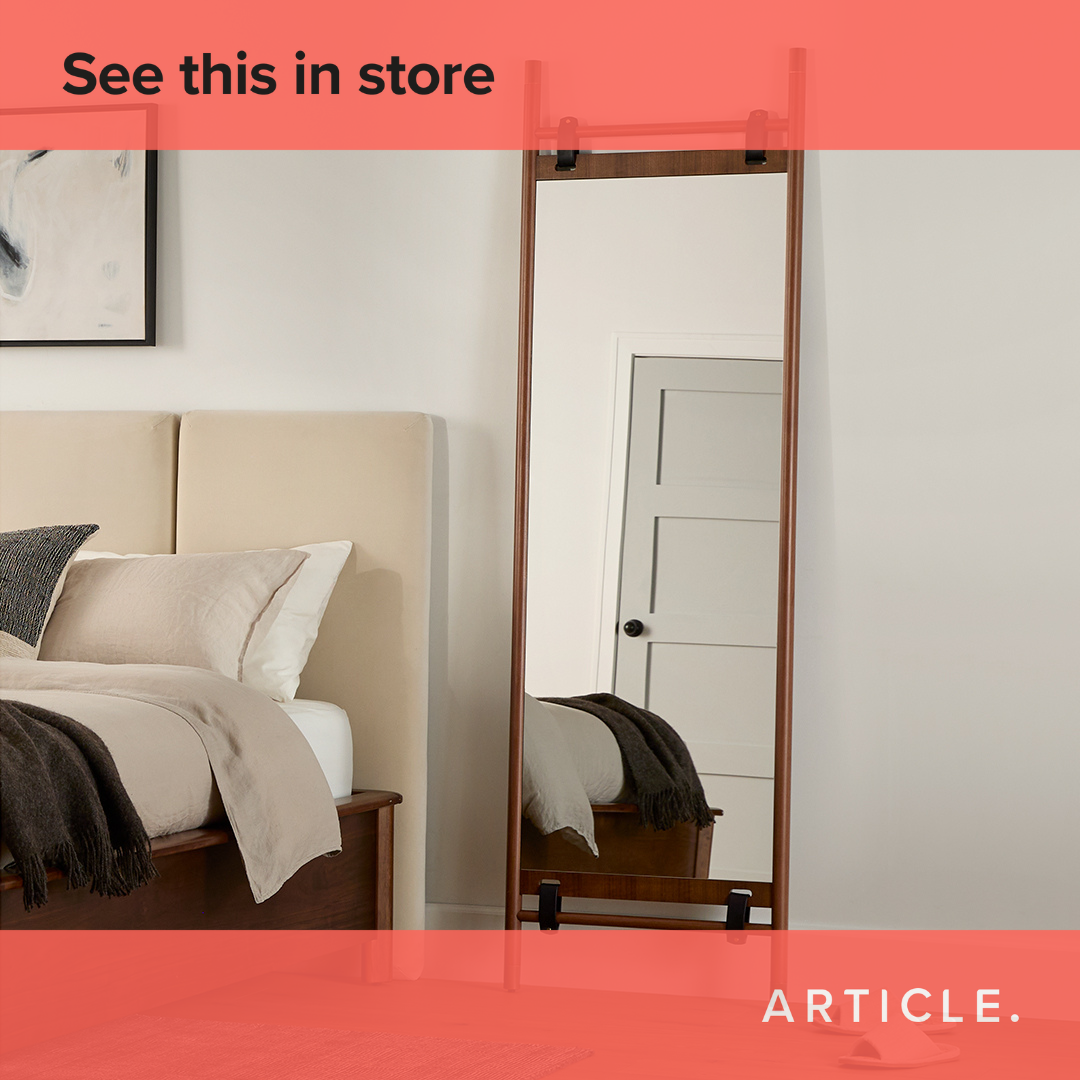 A full-length mirror with a wooden frame stands in a bedroom next to a bed with neutral-toned bedding. The mirror reflects a closed door and a portion of the bed. Text overlayed at the top of the image reads "See this in store". The brand name "ARTICLE." is visible in the bottom right corner.