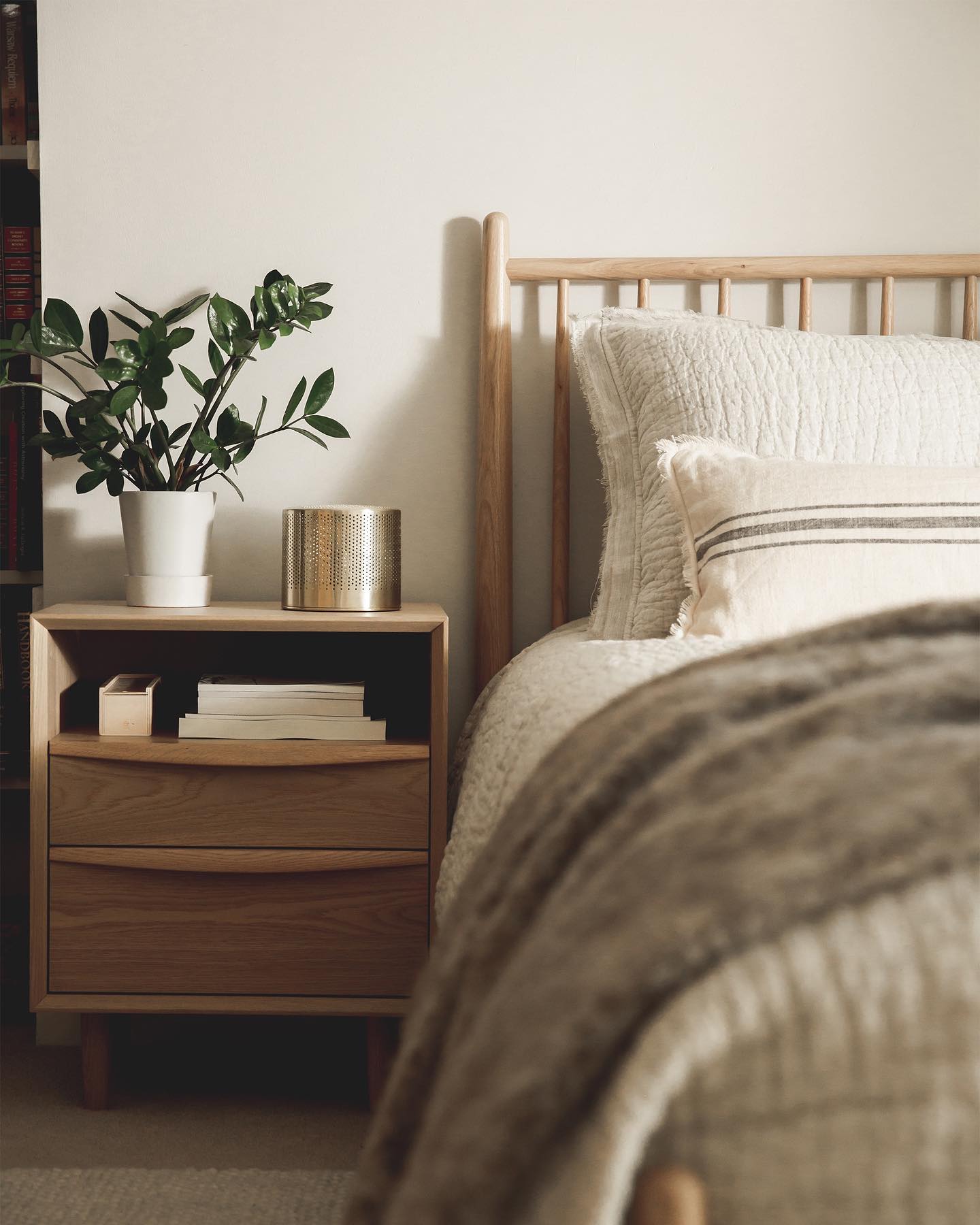 A close-up, softly lit shot of a neatly made bed with a wooden headboard. A bedside table made of light wood holds a potted plant and a decorative metallic cylinder. Books are stacked on the lower shelf of the nightstand. The overall aesthetic is minimalist and serene, suggesting a calm and comfortable bedroom environment. There is no text on the image.