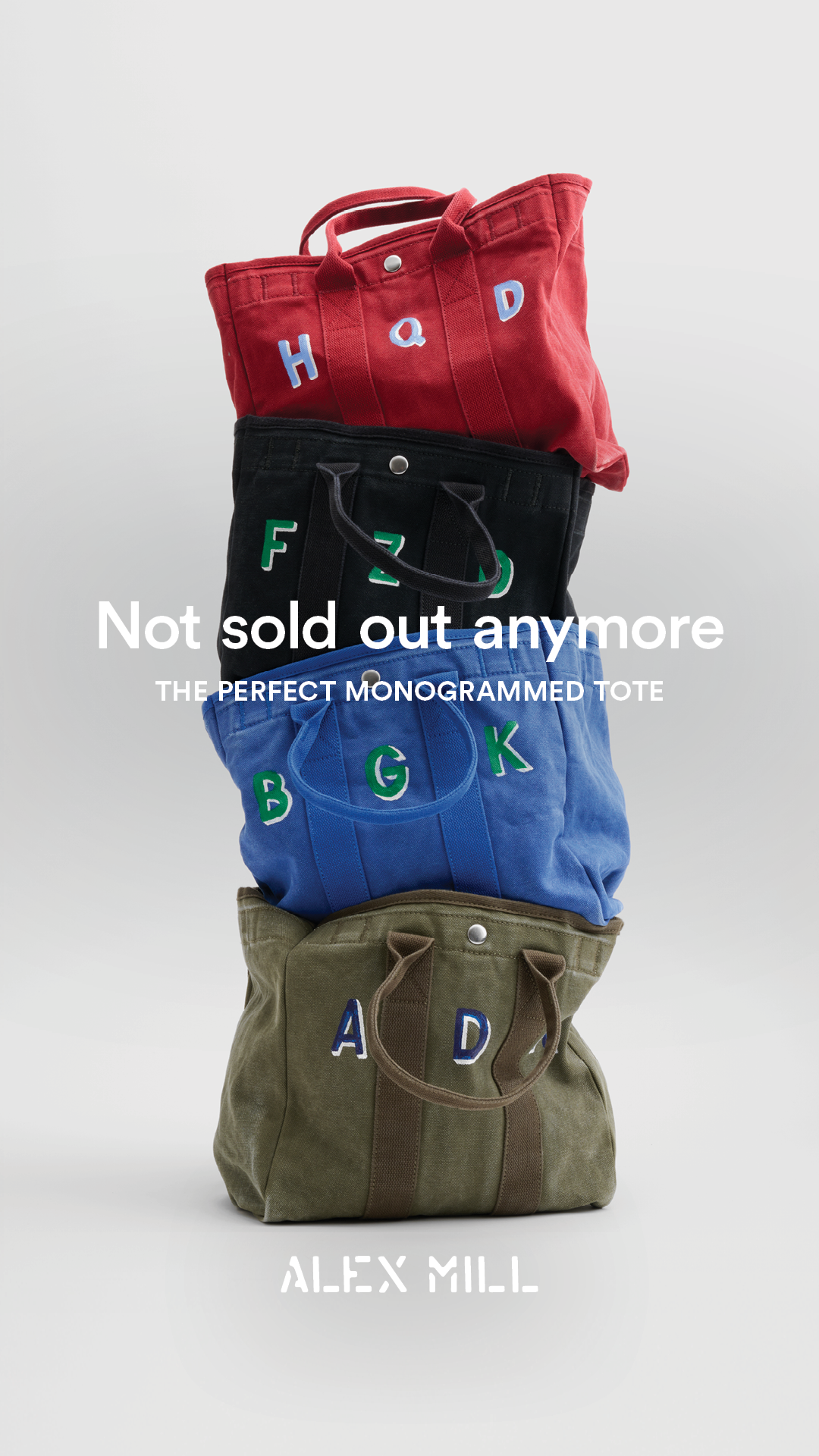 A stack of four canvas tote bags in different colors (red, black, blue, and olive green) are shown against a plain white background. Each bag has large, embroidered letters on the front, spelling out different words. Text overlays read "Not sold out anymore" and "THE PERFECT MONOGRAMMED TOTE". Below the bags, the brand name "ALEX MILL" is displayed in white stencil-like lettering. The creative highlights the return of a popular product that has previously sold out.