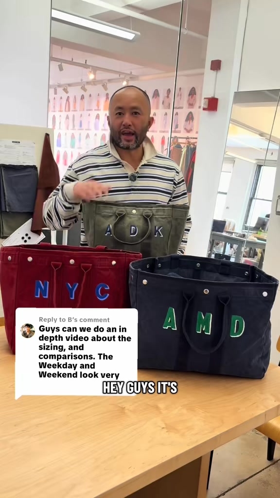 A man stands behind a table displaying three canvas tote bags in different colors: red, olive green, and navy blue. Each bag has large, embroidered letters on the front. A comment box is overlaid on the bottom left of the screen, with a user asking for an in-depth video about the sizing and comparisons of the bags. The man is gesturing towards the bags and speaking to the camera, with the text "HEY GUYS IT'S" overlaid on the bottom right of the screen.