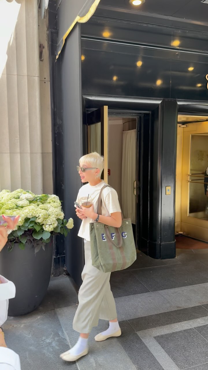 A young woman with short, blonde hair and sunglasses walks past a large planter filled with white hydrangeas. She is wearing a cream t-shirt, light-colored pants, and white socks with cream flats. She carries a green tote bag with the letters "EFC" embroidered on it and holds a to-go cup with a straw. The background shows the entrance to a building with black doors and gold accents. On-screen text announces a "BIG DEAL ALERT" and mentions that the woman, @emmachamberlain, was spotted with the brand's tote bag.