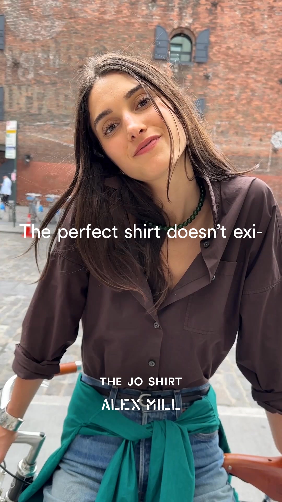 A young woman with long, dark hair is shown from the chest up, leaning slightly towards the camera with a gentle smile. She is wearing a dark brown button-up shirt and a green beaded necklace. The text "The perfect shirt doesn't exist-" is overlaid in white, sans-serif font across the middle of the image. Below that, "THE JO SHIRT" and "ALEX MILL" are also displayed in white text. The background appears to be an urban street scene with a brick building.