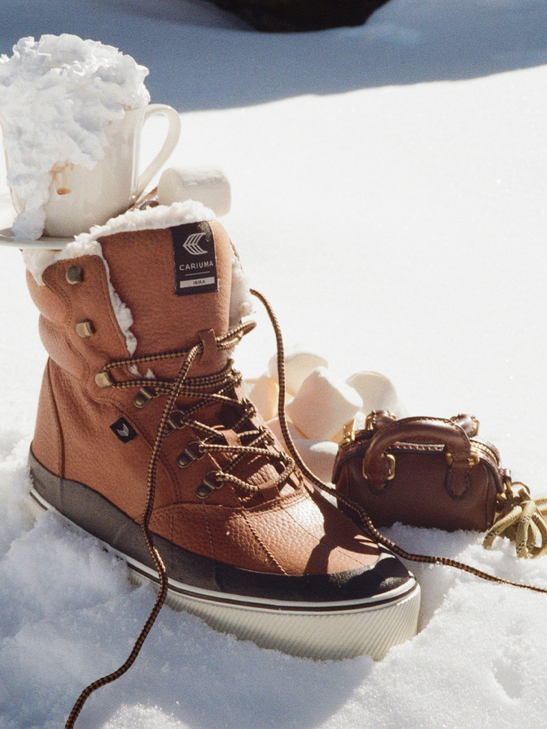A close-up, eye-level shot of a tan leather winter boot with white fur lining, sitting in the snow. A white mug overflowing with whipped cream and a hot beverage is perched on the boot's laces. To the right of the boot, a small, brown, structured handbag with a handle and tassel detail rests in the snow. The text overlay reads: "Zima si žádá teplo a jistotu v každém kroku." (Winter demands warmth and confidence in every step.)