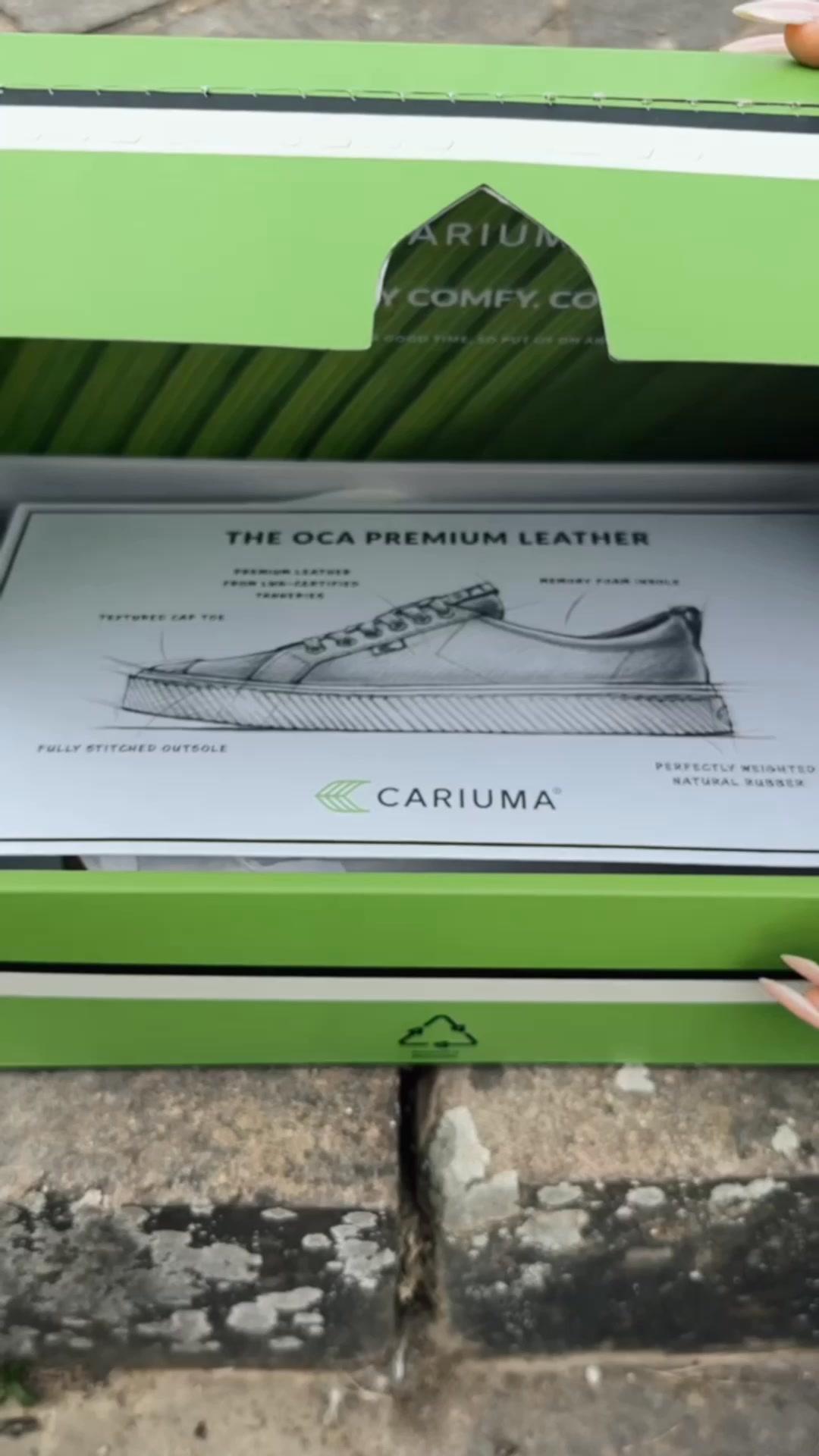 The video opens with a shot of a green Cariuma shoebox being opened. Inside, a detailed sketch of a Cariuma sneaker is displayed on a white card. The sketch highlights various features of the shoe, such as 'PREMIUM LEATHER', 'TEXTURED CAP TOE', 'FULLY STITCHED OUTSOLE', 'MEMORY FOAM INSOLE', and 'PERFECTLY WEIGHTED NATURAL RUBBER'. The Cariuma logo is also visible on the card. The text on the screen reads: 'Cariuma.de – Diese brasilianischen Schuhe sind ein weltweiter Hit! Getragen von Top-Skatern und Hollywood-Stars. Extrem bequem. Unglaublich strapazierfähig. Nachhaltig hergestellt. In vielen Farben, passend zu Ihrem Stil. Für jedes Paar pflanzen wir 2 Bäume. Der Versand ist kostenlos. Sie erhalten 20 € Rabatt auf Ihren ersten Einkauf. Und falls sie nicht passen sollten, können Sie sie ganz einfach zurückgeben.'