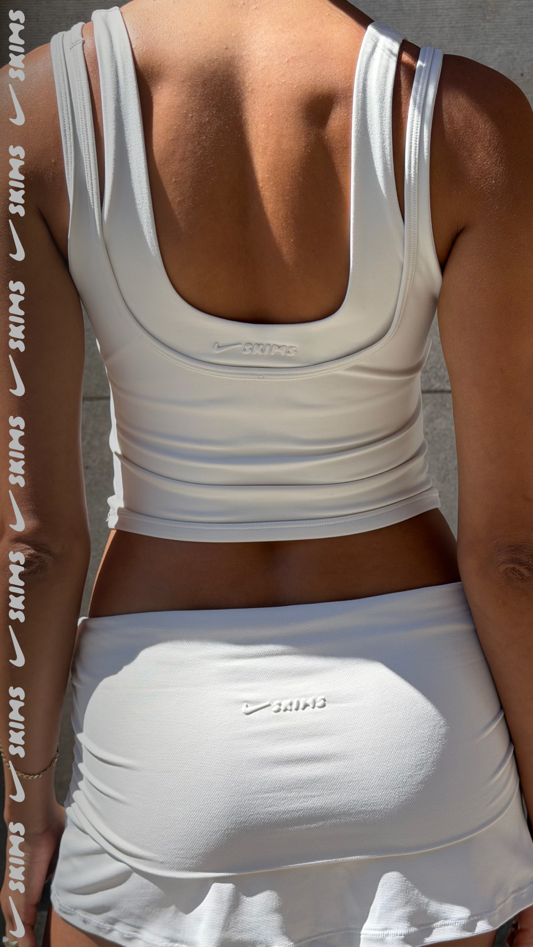 The back of a person wearing a white Skims tank top and skirt. The Skims logo is visible on both the tank top and the skirt. White text with Nike swoosh logos is overlaid vertically on the left side of the image, reading "SKIMS" repeatedly. The hook text, "A studio essential you can wear anywhere. Shop our best selling Matte Double Strap Scoop Tank with Nike Dri-FIT™ technology!", is also overlaid on the image.