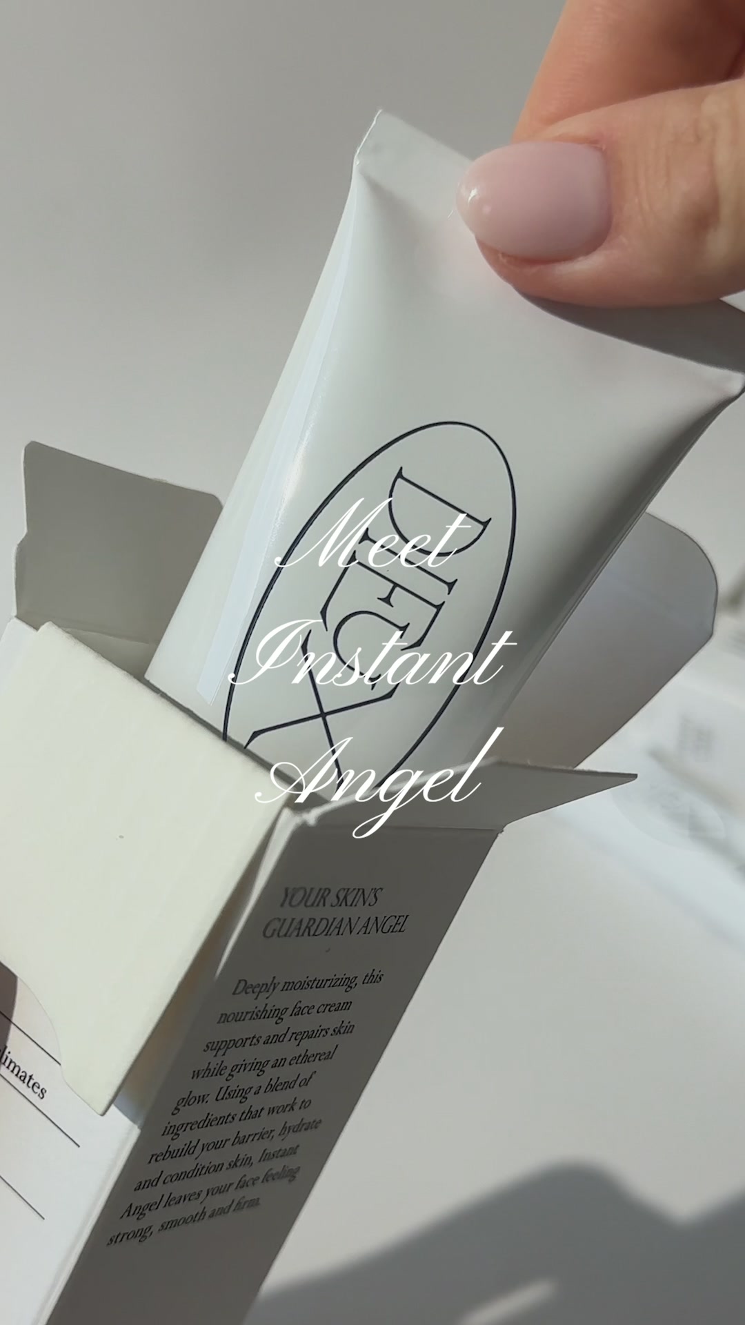 A hand with a light pink manicure is holding a white tube of face cream, pulling it out of its open white box. The text "Meet Instant Angel" is overlaid in a script font on the cream tube and box. Below that, smaller text reads "YOUR SKINS GUARDIAN ANGEL" and then a paragraph describing the product's benefits. The overall aesthetic is clean and minimalist, focusing on the product itself.