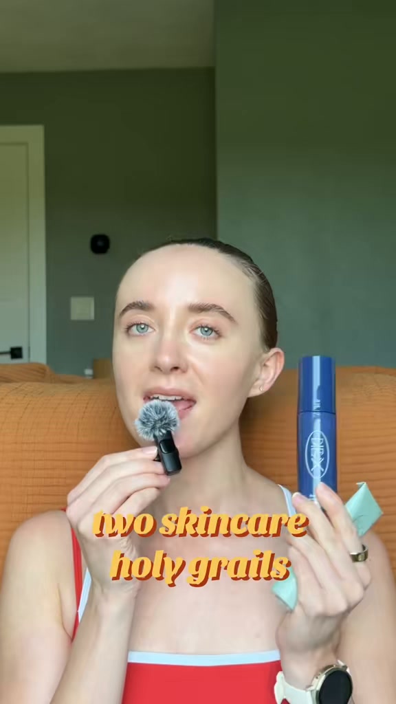 A woman with her hair pulled back is holding a small microphone near her mouth and a blue bottle of skincare product in her other hand. Yellow text overlaid on the video reads "two skincare holy grails." The woman is wearing a red and white striped top. The background is a muted green wall and an orange textured couch. The creative appears to be a personal recommendation or review of skincare products.
