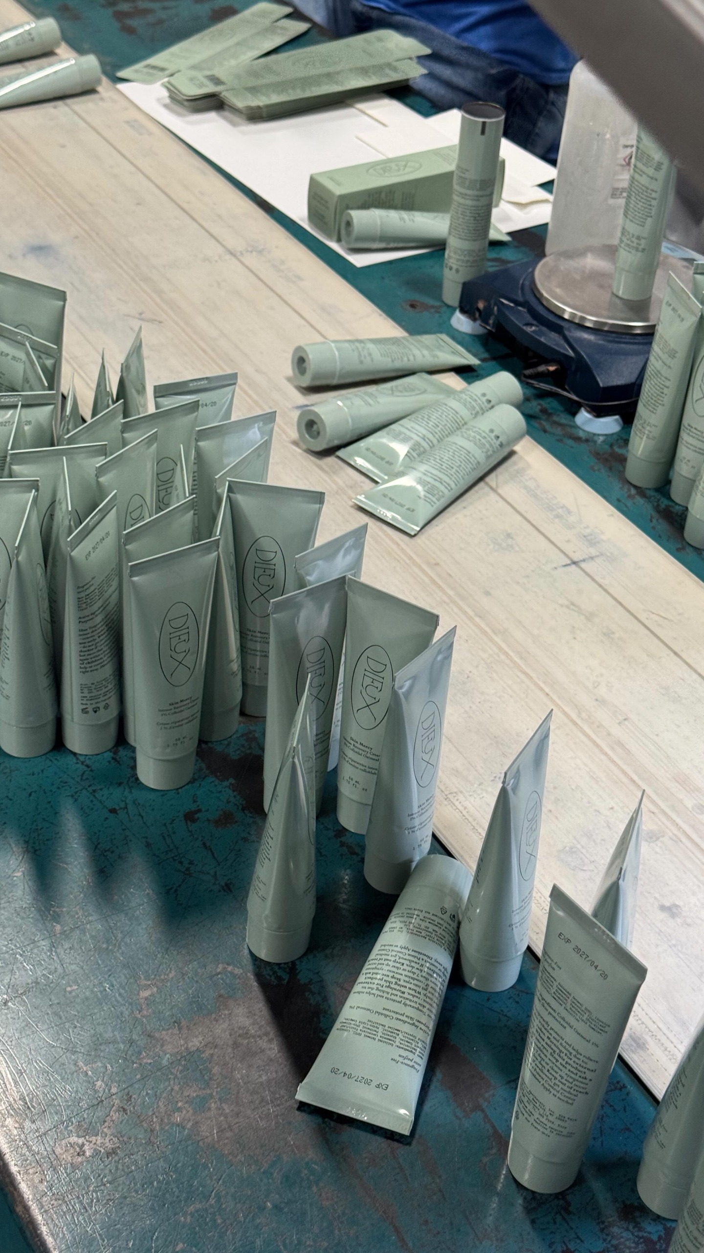 The image shows a close-up, overhead view of numerous tubes of skincare products arranged on a wooden surface. Some tubes are standing upright in rows, while others are lying down. The tubes are a pale green color with minimalist branding. In the background, there are stacks of boxes and a digital scale with a bottle and a tube on it, suggesting a production or packaging environment. The overall impression is one of meticulous preparation and scientific rigor in the creation of these products.