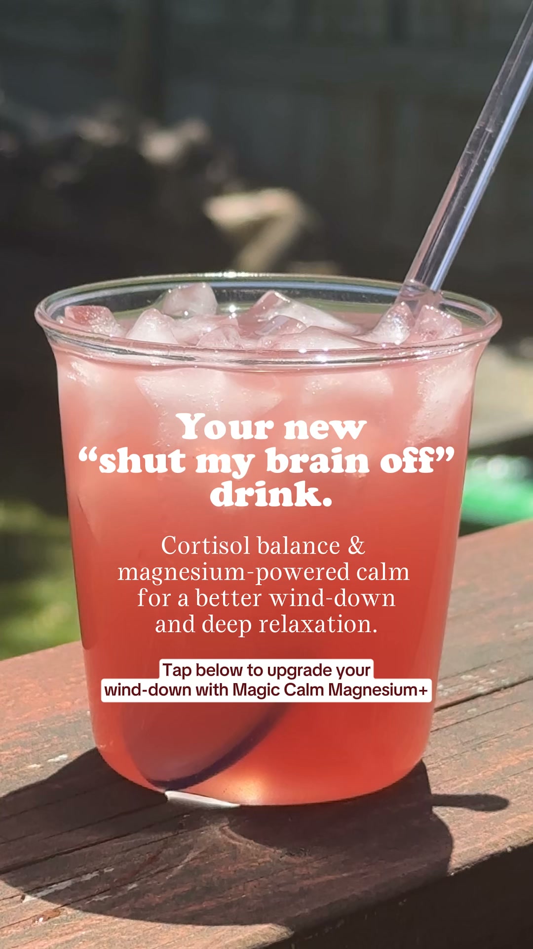 A close-up shot of a pink, iced beverage in a clear plastic cup, sitting on a wooden surface outdoors. White text overlays the drink, with the main headline reading "Your new \"shut my brain off\" drink." Below that, smaller text explains "Cortisol balance & magnesium-powered calm for a better wind-down and deep relaxation." At the bottom, a call to action states, "Tap below to upgrade your wind-down with Magic Calm Magnesium+."