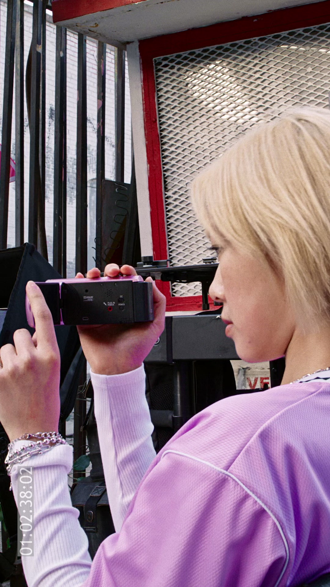 A close-up shot shows a person holding a pink camcorder, looking towards the right side of the frame. The person is wearing a purple long-sleeved shirt over a white ribbed undershirt and has several silver bracelets on their left wrist. The background features a metal fence and a red-trimmed structure. Text overlaying the image reads: "The iconic Saucony Omni 9 just keeps on evolving."