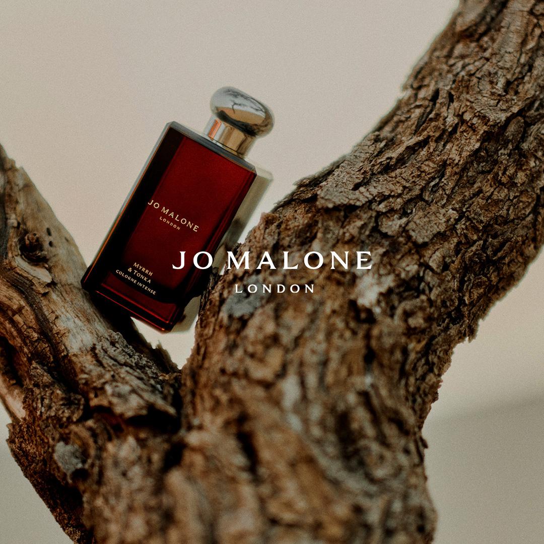 A dark red Jo Malone London perfume bottle rests on a rough, textured tree branch. The bottle is angled, with its silver cap glinting. The brand name and product details are visible on the bottle. The background is a soft, out-of-focus light color. Text overlays the image, featuring the brand name and a descriptive hook about the scent evoking desert dunes and pure elegance.
