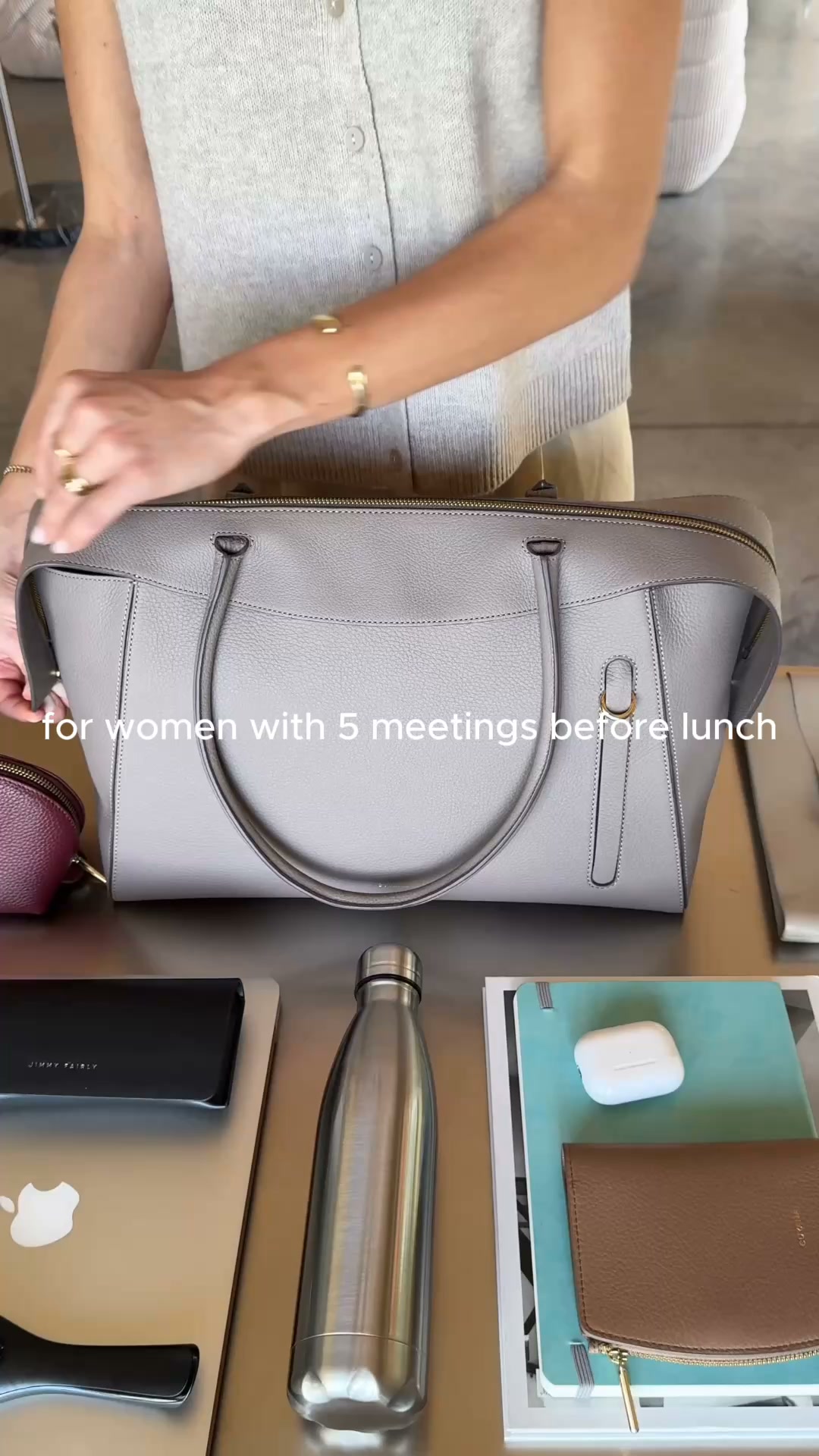 A woman's hands are shown arranging items on a table next to a large, light grey leather tote bag. The text overlay reads "for women with 5 meetings before lunch". On the table are a laptop, a metal water bottle, a notebook, wireless earbuds, and a wallet. The scene depicts a woman preparing her bag for a busy day of professional engagements.