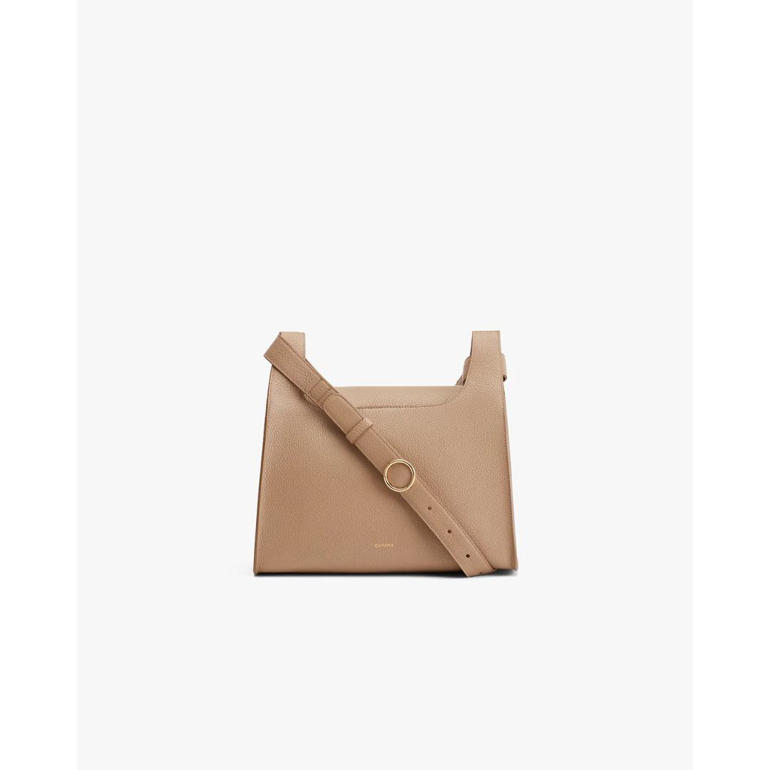 A clean, studio shot of a light brown, pebbled leather crossbody bag is centered against a plain white background. The bag has a structured, rectangular shape with a flap closure and a single adjustable strap that goes across the front. The strap is secured with a gold-toned circular buckle. The brand name 'CUYANA' is subtly embossed on the lower front of the bag. The overall aesthetic is minimalist and elegant, highlighting the craftsmanship and material of the accessory.