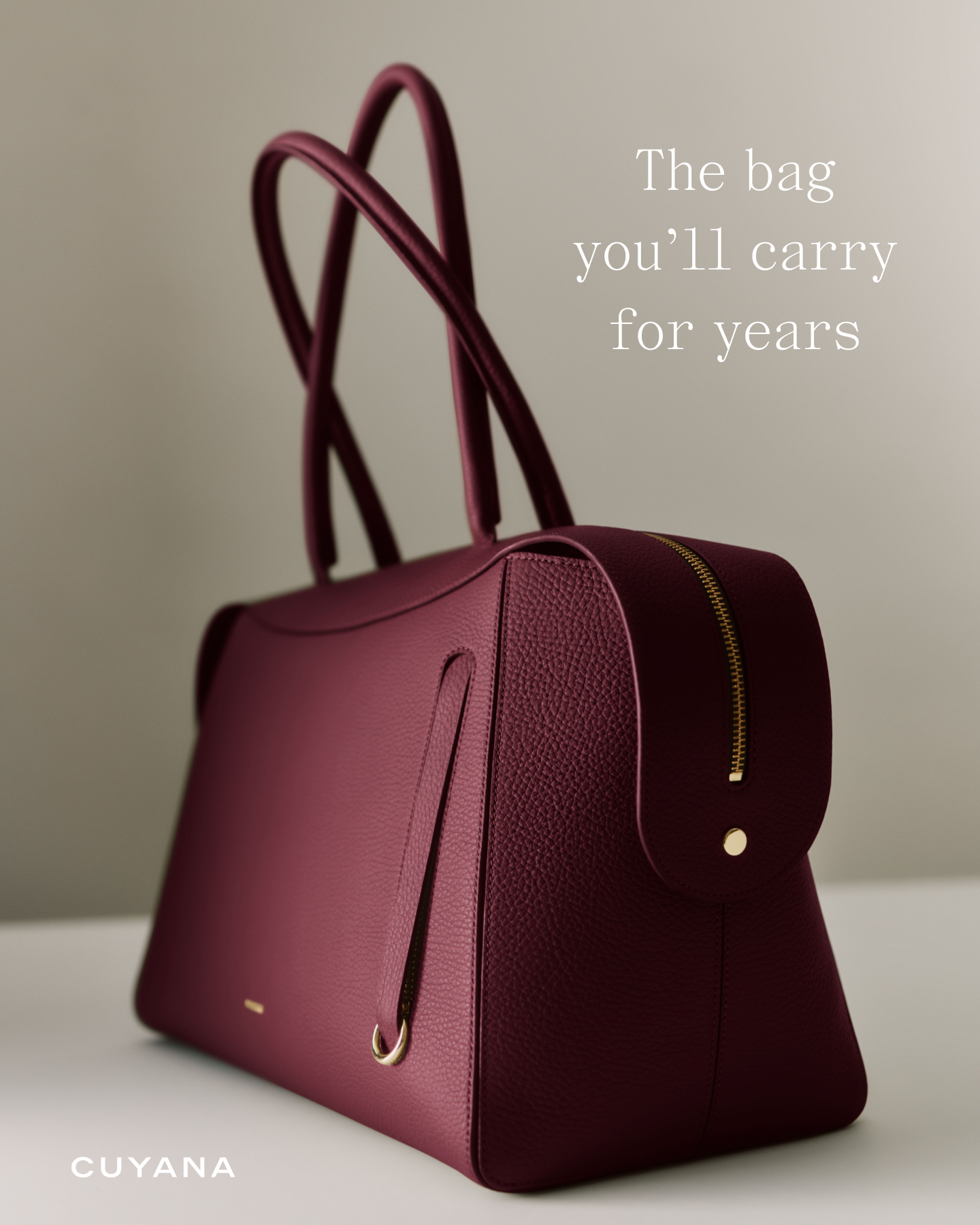 A deep burgundy handbag is shown from a low angle, placed on a white surface against a neutral grey background. The bag has a textured leather finish and gold hardware, including a prominent zipper. White text overlays the image, reading "The bag you'll carry for years" in a serif font. The brand name "CUYANA" is visible in white text at the bottom left corner of the image.