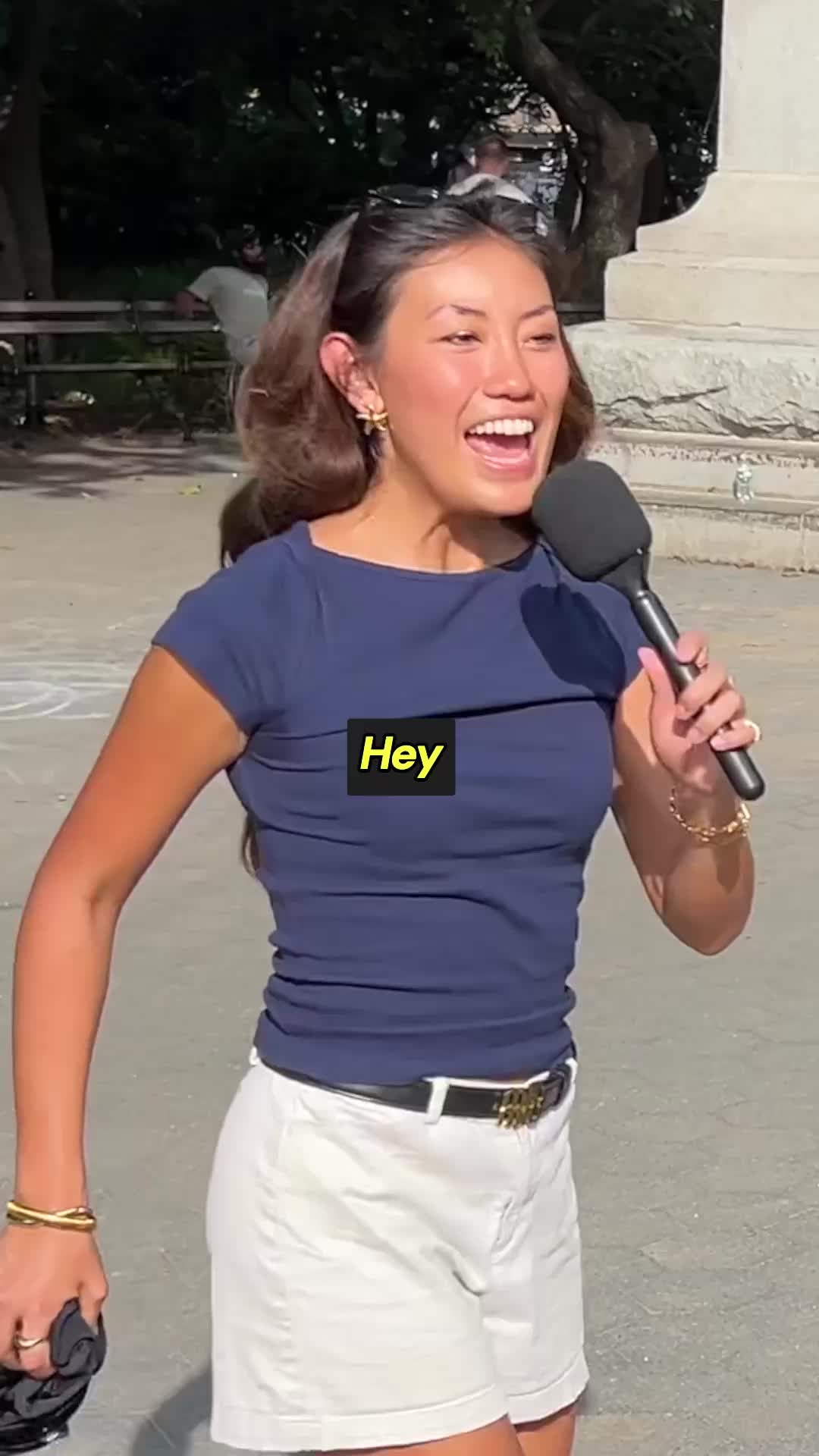 A woman with dark hair pulled back in ponytails, wearing a navy blue t-shirt and white shorts, is holding a microphone and speaking outdoors. A yellow text box with the word "Hey" is overlaid on her shirt. She appears to be in the middle of a street interview, with a blurred background of trees and a stone structure. The overall impression is of a casual, engaging interaction.