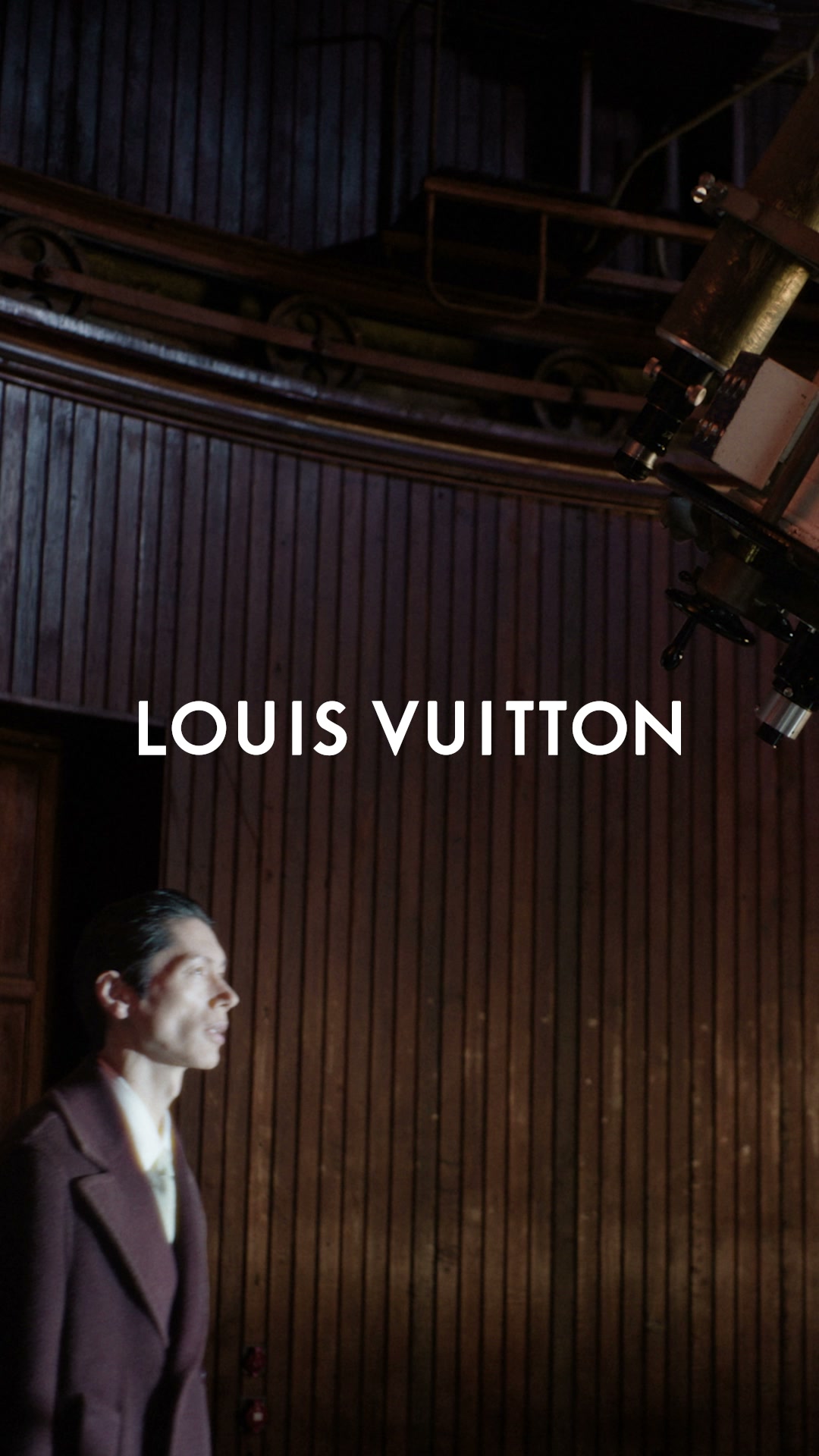 A man in a dark suit stands in a room with dark wood-paneled walls, looking upwards towards a large telescope. The Louis Vuitton logo is displayed prominently in white text across the center of the image. The text overlay reads 'Autunno-Inverno 2026 Spinta da aspirazioni cosmiche, la nuova collezione di Pharrell Williams svela un guardaroba tecnico ed elegante.' This suggests a collection inspired by cosmic aspirations and featuring technical, elegant apparel.