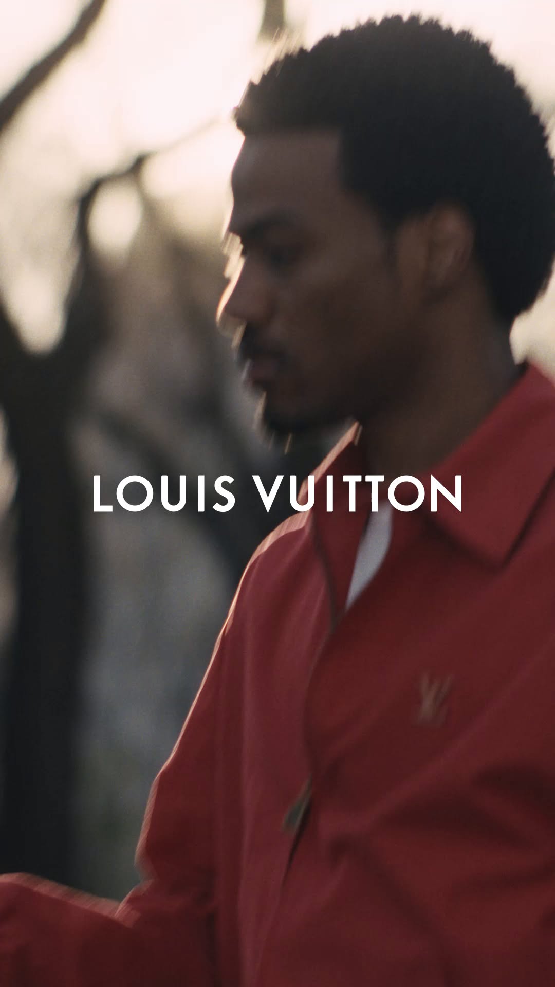 A close-up, profile shot of a Black man with an afro hairstyle, wearing a red collared shirt. The Louis Vuitton logo is visible on the shirt. White text overlays the image, reading "LOUIS VUITTON". The background is blurred, suggesting an outdoor setting with soft, natural light. The man appears to be looking off to the side, with a thoughtful expression. The overall aesthetic is sophisticated and modern, highlighting the brand and the individual.