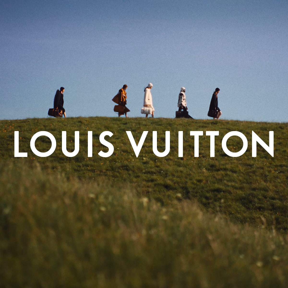 A group of five people walk single file across a grassy hill under a clear blue sky. They are dressed in elegant, layered clothing, suggesting a fashion context. The Louis Vuitton logo is prominently displayed in large white sans-serif text across the lower portion of the image. The copy explains that the collection is inspired by Jura, the birthplace of the founder, Louis Vuitton, and highlights Nicolas Ghesquière's latest collection.