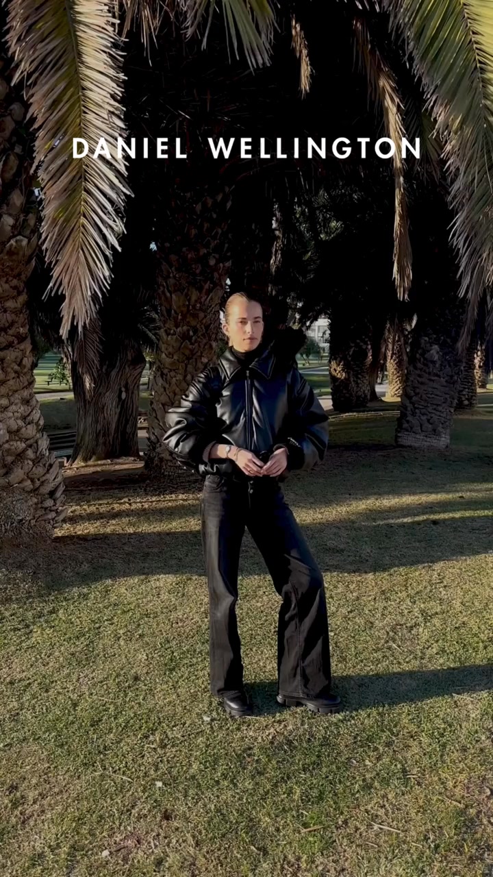 A person stands in a park with palm trees in the background. They are wearing a black leather jacket and black pants. The text "DANIEL WELLINGTON" is displayed at the top of the image. The hook "La sicurezza è nei dettagli." is presented, followed by "Realizzato alla perfezione." which translates to "Made to perfection." The overall aesthetic is chic and sophisticated, highlighting the brand's attention to detail in its products.
