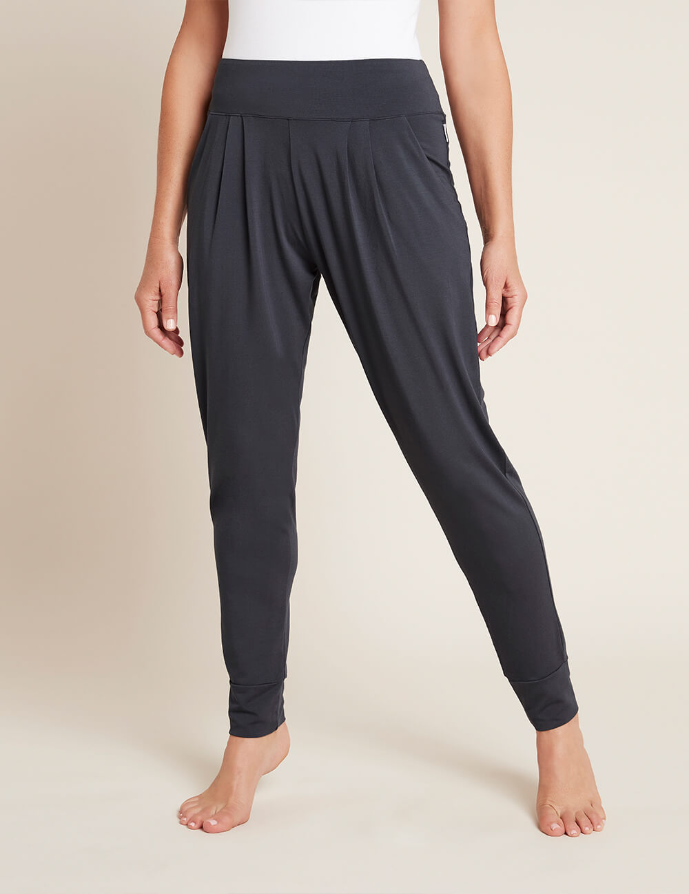 A medium shot shows the lower body of a woman wearing dark grey, loose-fitting, high-waisted pants made of a soft, flowing fabric. The pants have pleats at the front and cuffed ankles. She is barefoot, standing on a plain, light-colored surface against a similar background. The text "Consciously crafted in organically-grown bamboo viscose, our products help you live a little easier, and a lot comfier." is overlaid on the image, followed by "Over 120,000 5-star reviews ⭐⭐⭐⭐⭐".