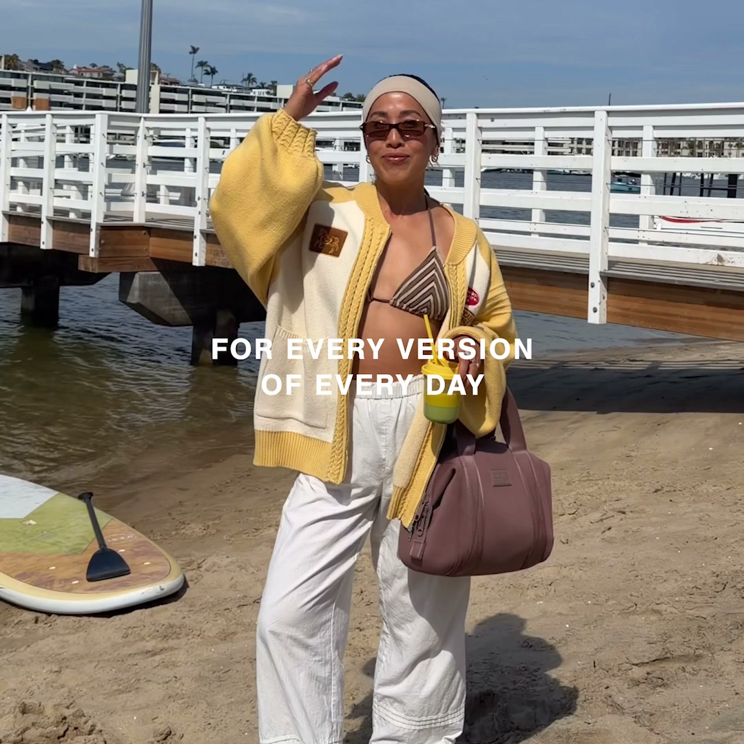A woman stands on a sandy beach, wearing a yellow cardigan over a bikini top and white pants. She holds a drink in her right hand and has her left hand raised to her forehead, shielding her eyes from the sun. Behind her, a white pier extends over the water, and a paddleboard rests on the sand to her left. White text overlaid on the image reads: "FOR EVERY VERSION OF EVERY DAY."