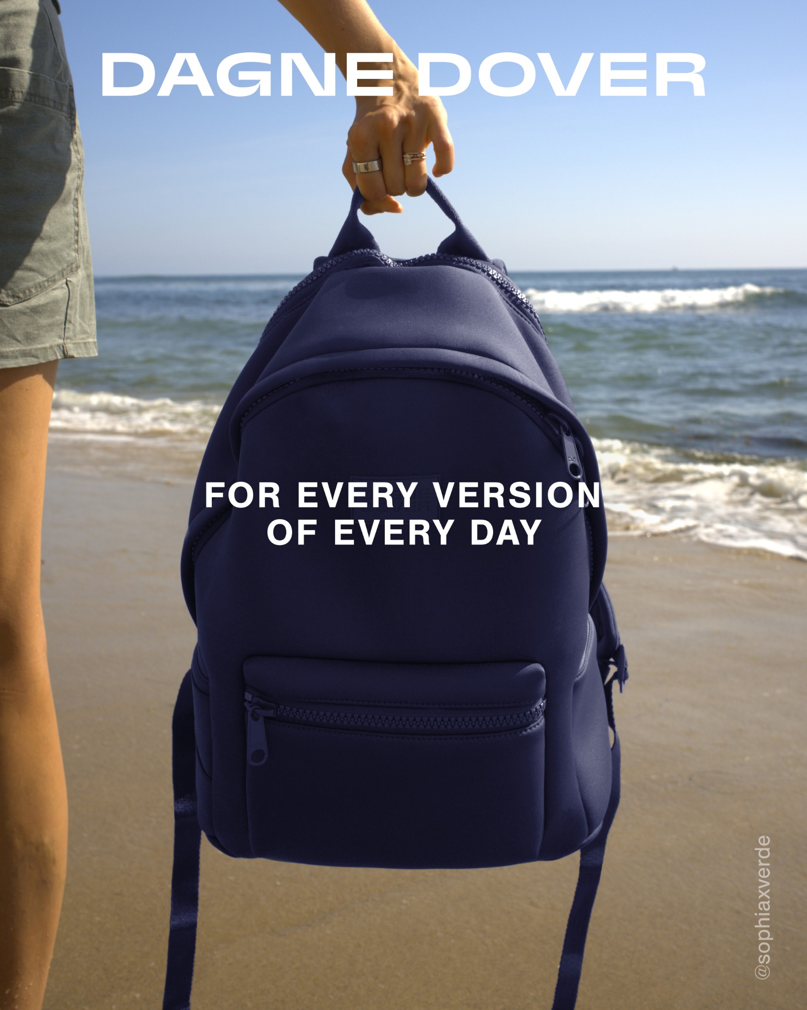 A person stands on a beach, holding a navy blue backpack by its straps. The ocean and waves are visible in the background under a clear blue sky. White text overlaid on the image reads "DAGNE DOVER" at the top and "FOR EVERY VERSION OF EVERY DAY" across the backpack. The overall scene evokes a sense of everyday use and versatility for the product.
