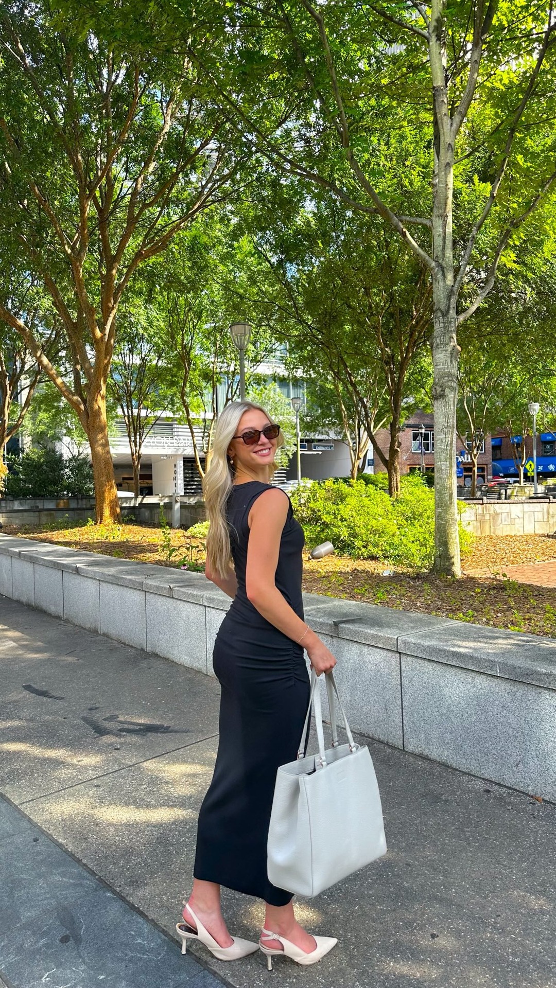 A woman with blonde hair, wearing sunglasses and a black sleeveless dress, smiles over her shoulder while walking outdoors. She carries a large, light-colored tote bag. The background features trees and a building. Text overlays the image, stating "I love my Dagne Dover bag for work and workouts!"